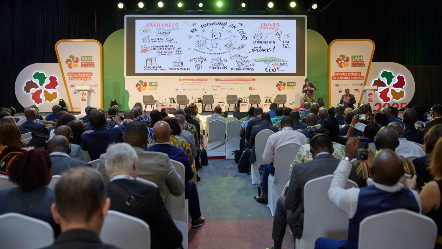 Une foule assise sur des chaises blanches devant un écran, vue de dos